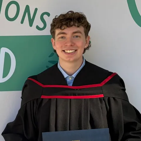 Cole at graduation, smiling in a black gown and holding a UBC diploma folder